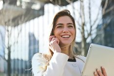 Femme heureuse, tient un ordinateur en main et est au téléphone, télétravailleur en extérieur, elle est épanouit